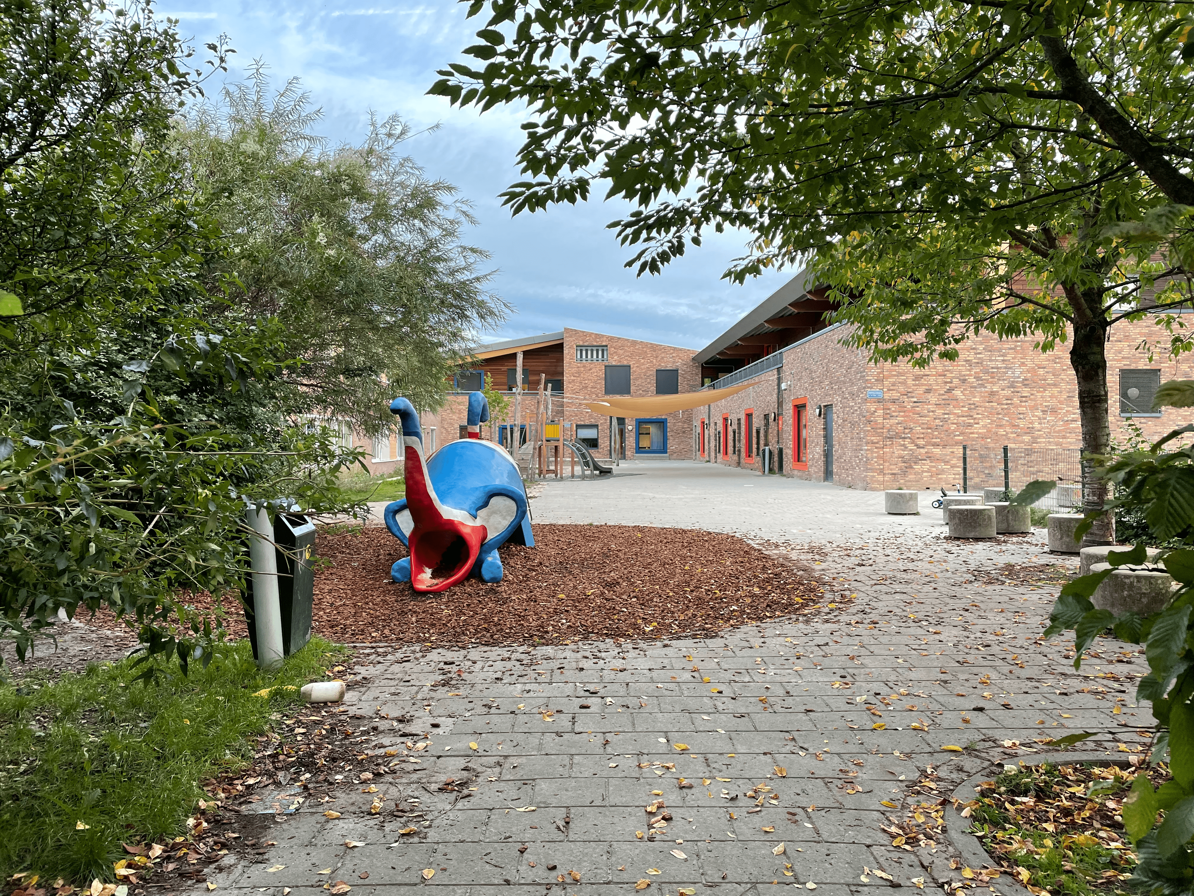 Schoolplein Bunnik met een speeltoestel in de vorm van een blauw, fictief dier. Op de achtergrond het U-vormige schoolgebouw om het schoolplein heen.